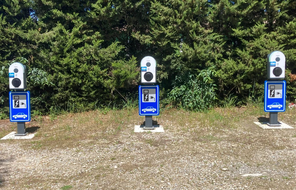 Trois bornes de recharge pour véhicules électriques installées en plein air sur un parking graveleux, fond végétal dense