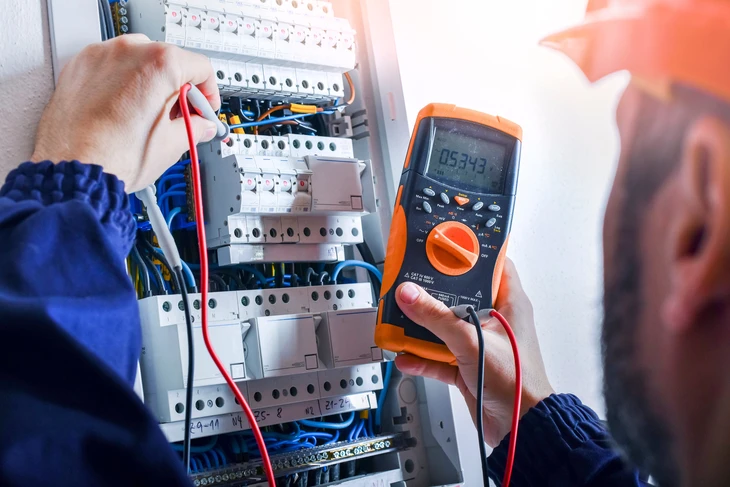 Photographie en intérieur d'un électricien professionnel équipé d'outils travaillant sur un tableau électrique ou une installation électrique murale, montrant une intervention calme et professionnelle. L'électricien porte des équipements de sécurité (gants, vêtements professionnels). L'environnement est un intérieur domestique clair et bien éclairé, sans danger apparent. La scène transmet confiance, professionnalisme et compétence dans la résolution de problèmes électriques.