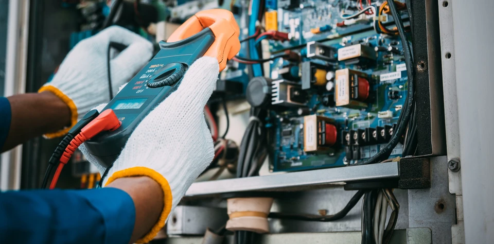 Technicien en combinaison bleue effectuant des mesures électriques sur un tableau de contrôle industriel avec un multimètre à pinces ampèremétriques