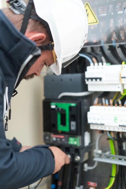 Électricien en uniforme bleu travaillant dans un tableau électrique industriel, utilisant une pince ampèremétrique orange pour mesurer l'intensité électrique